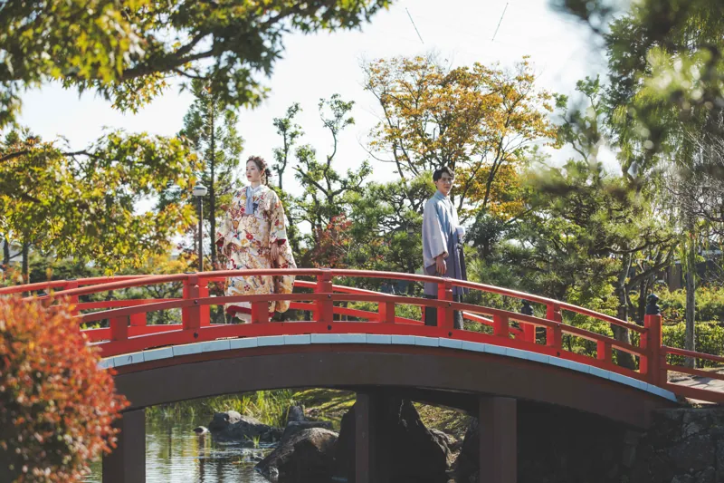 紫式部公園の朱色の橋の上に立つ、色打掛と羽織袴のおふたり