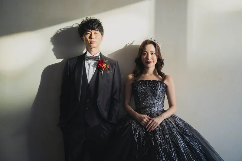 光の差しこむ白い壁の前に並ぶ、紺色のドレスの花嫁とタキシードの新郎