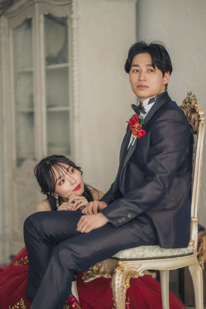 赤いドレスの花嫁が、アンティークの椅子に座る新郎にもたれかかる様子